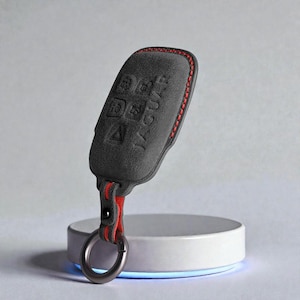 以下が含まれることがあります： 赤いステッチと赤いストラップが付いた、濃い灰色のキーフォブカバー。カバーにはエンボス加工されたボタンと「JAGUAR」の文字があります。シルバーカラーのキーリングがストラップに付いています。キーフォブは白い円形のスタンドに置かれています。