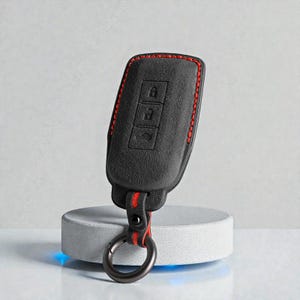 Puede incluir: Funda negra para llavero de coche con costuras y detalles en rojo. La funda tiene tres botones y un llavero de metal. El llavero se muestra sobre un soporte cilíndrico blanco sobre un fondo gris claro.
