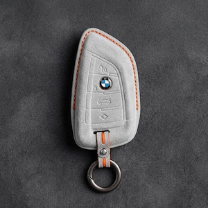 以下が含まれることがあります： BMW車用ライトグレーのスエード製キーフォブカバー。縁にはオレンジ色のコントラストステッチが施されています。カバーにはロック、アンロック、トランク、パニック機能のエンボス加工されたアイコンがあり、ベース部分には金属製のリングが取り付けられています。