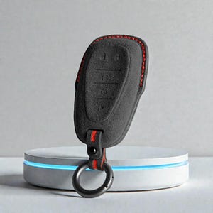 May include: A black car key fob cover with red stitching and a red and black strap. The key fob cover has buttons for locking, unlocking, and other functions. A black metal ring is attached to the strap.