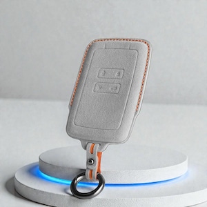 Könnte beinhalten: Eine graue Autoschlüsselhülle mit orangefarbenen Nähten und einem passenden orangefarbenen Riemen. Die Schlüsselhülle hat einen schwarzen Metallring und wird auf einem weißen Sockel mit blauem Licht präsentiert.