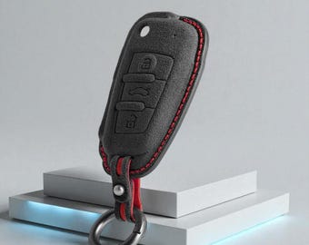 Könnte beinhalten: Eine schwarze Autoschlüsselhülle mit roten Nähten und einem rot-schwarzen Riemen. Die Hülle hat drei Knöpfe und einen Metallschlüsselring. Der Schlüsselanhänger liegt auf einer weißen Oberfläche.