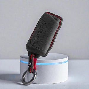 Può includere: Una custodia nera per portachiavi con cuciture rosse e un cinturino rosso. Il portachiavi ha dei pulsanti e un anello di metallo per attaccare le chiavi. La custodia è realizzata in un materiale strutturato e viene esposta su un piedistallo bianco.