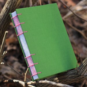 Puede incluir: Un diario hecho a mano con una cubierta verde brillante y costuras rosas. Las p&aacute;ginas son blancas y la encuadernaci&oacute;n es visible a lo largo del lomo. El diario descansa sobre ramas marrones, creando un fondo natural.