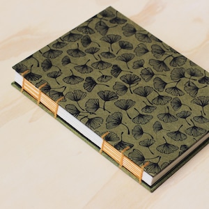 Könnte beinhalten: Ein handgefertigtes Notizbuch mit grünem Einband und einem schwarzen Ginkgo-Blattmuster. Die Seiten sind mit orangefarbenem Faden gebunden, und die Kanten sind grün. Das Notizbuch ist geschlossen und liegt auf einer hellen Holzoberfläche.