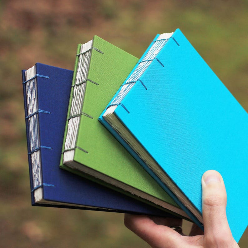Puede incluir: Tres cuadernos hechos a mano en tonos azul, verde y turquesa. Las cubiertas son de color liso y las p&aacute;ginas est&aacute;n encuadernadas con costuras visibles. Los cuadernos se sostienen en una mano, mostrando su tama&ntilde;o y dise&ntilde;o.