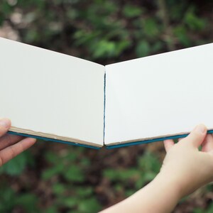 Puede incluir: Un cuaderno de bocetos abierto con p&aacute;ginas blancas en blanco, sostenido contra un fondo verde borroso. El libro tiene una cubierta azul y las p&aacute;ginas tienen un acabado texturizado. La imagen est&aacute; bien iluminada, destacando la textura del papel y el dise&ntilde;o del libro.