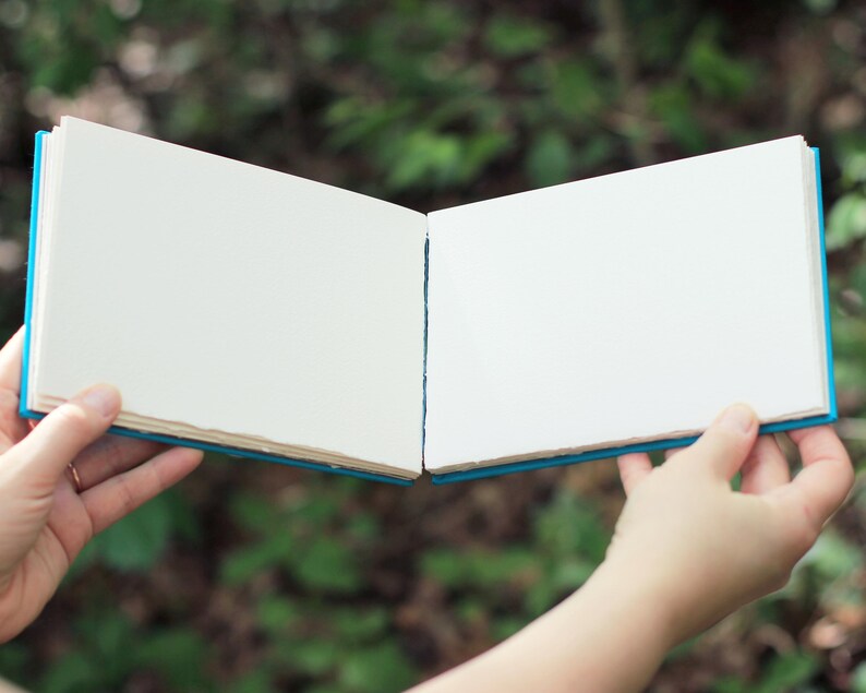 Puede incluir: Un cuaderno de bocetos abierto con p&aacute;ginas blancas en blanco, sostenido contra un fondo verde borroso. El libro tiene una cubierta azul y las p&aacute;ginas tienen un acabado texturizado. La imagen est&aacute; bien iluminada, destacando la textura del papel y el dise&ntilde;o del libro.