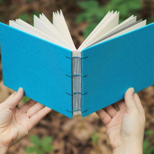 Puede incluir: Un diario hecho a mano con una cubierta azul turquesa brillante y encuadernaci&oacute;n expuesta. Las p&aacute;ginas son una mezcla de blanco y gris. El diario se sostiene abierto, mostrando el lomo cosido y las p&aacute;ginas desplegadas.