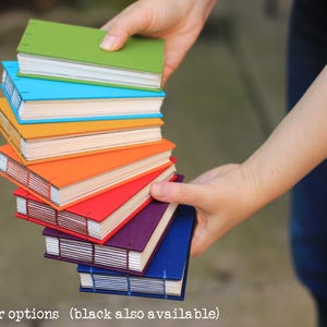 Puede incluir: Una pila de cuadernos hechos a mano y coloridos en varios tonos, incluyendo verde, azul, naranja, rojo, morado y azul. Los cuadernos son sostenidos en las manos de una persona, mostrando sus vibrantes cubiertas y encuadernaci&oacute;n cosida. El texto "color options (black also available)" est&aacute; en la parte inferior.
