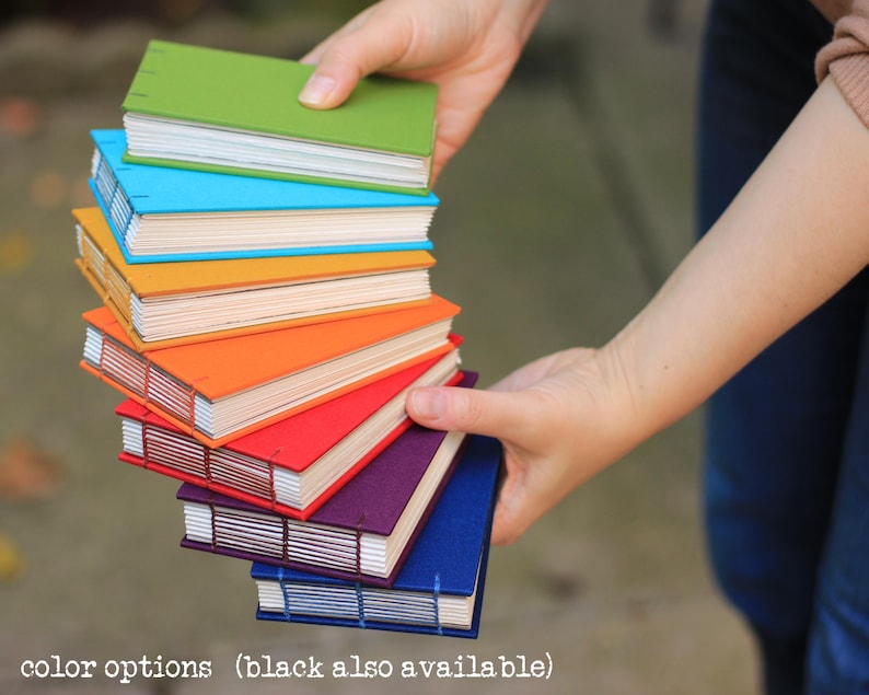 Puede incluir: Una pila de cuadernos hechos a mano y coloridos en varios tonos, incluyendo verde, azul, naranja, rojo, morado y azul. Los cuadernos son sostenidos en las manos de una persona, mostrando sus vibrantes cubiertas y encuadernaci&oacute;n cosida. El texto "color options (black also available)" est&aacute; en la parte inferior.