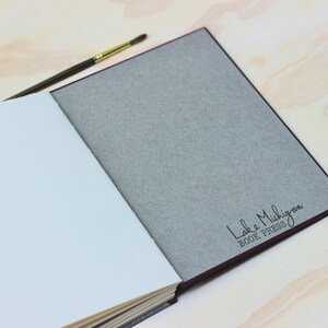 Puede incluir: Un libro abierto con p&aacute;ginas blancas y una cubierta gris. La cubierta tiene el texto "Lake Michigan BOOK PRESS" en escritura negra. Un pincel con mango marr&oacute;n y virola dorada descansa sobre el libro.