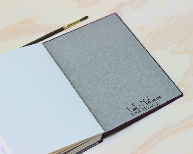 Puede incluir: Un libro abierto con p&aacute;ginas blancas y una cubierta gris. La cubierta tiene el texto "Lake Michigan BOOK PRESS" en escritura negra. Un pincel con mango marr&oacute;n y virola dorada descansa sobre el libro.