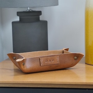 May include: A brown leather catchall tray with the word "DAD" embossed on the front. The tray is sitting on a wooden surface.