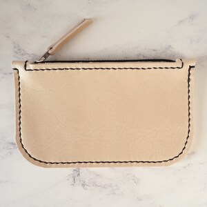 May include: A small, beige leather zipper pouch with black stitching. The zipper is open and the pouch is lying flat on a white marble surface.