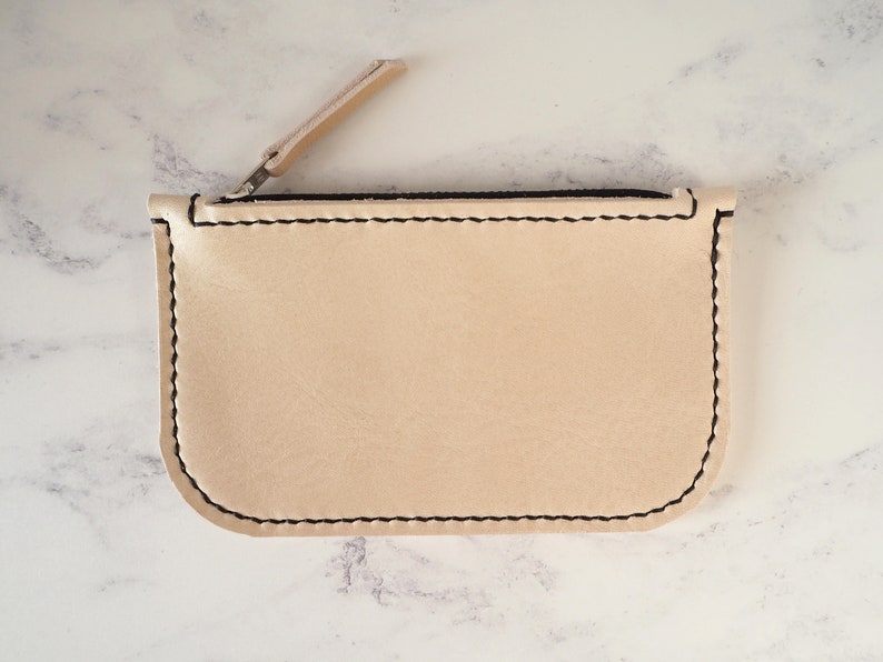 May include: A small, beige leather zipper pouch with black stitching. The zipper is open and the pouch is lying flat on a white marble surface.