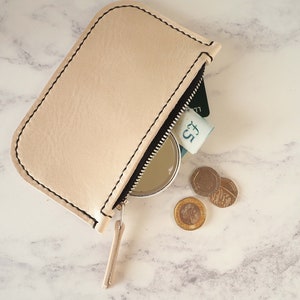 May include: A light beige leather zippered pouch with a black zipper pull. The pouch is open and contains a credit card, a five pound note, a small round mirror, and three British coins.