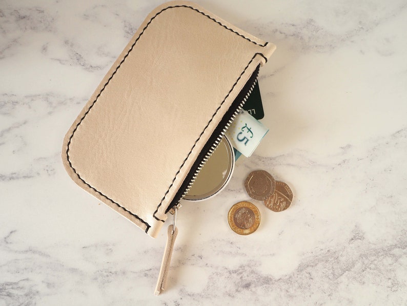 May include: A light beige leather zippered pouch with a black zipper pull. The pouch is open and contains a credit card, a five pound note, a small round mirror, and three British coins.