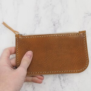 May include: A brown leather zippered pouch with white stitching. The pouch is held in a hand.
