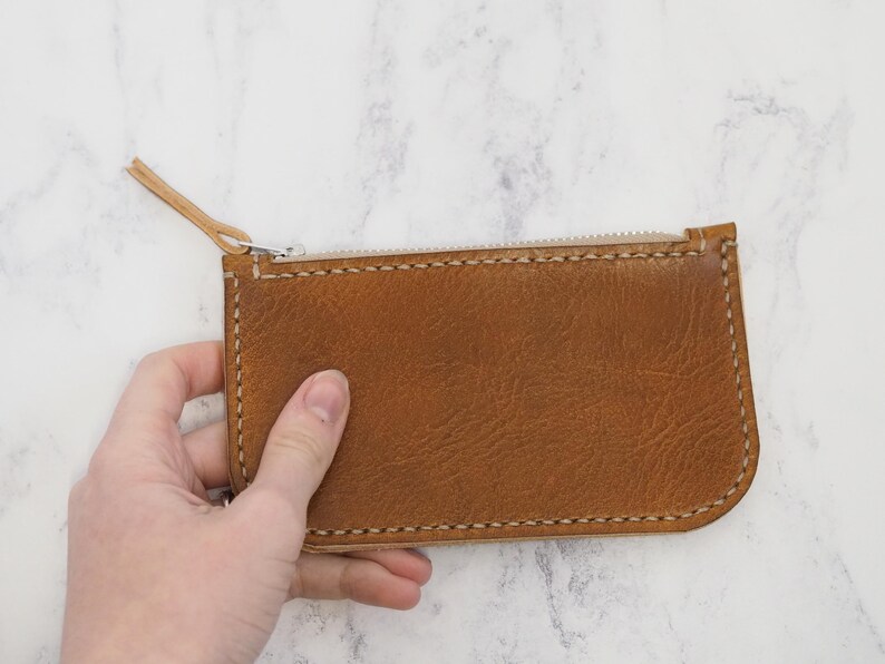 May include: A brown leather zippered pouch with white stitching. The pouch is held in a hand.