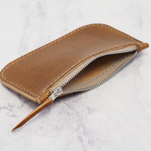 May include: A brown leather zipper pouch with a zipper pull. The pouch is open and the inside is lined with a light brown fabric.