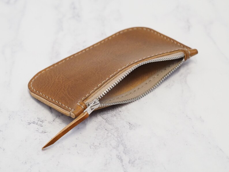 May include: A brown leather zipper pouch with a zipper pull. The pouch is open and the inside is lined with a light brown fabric.