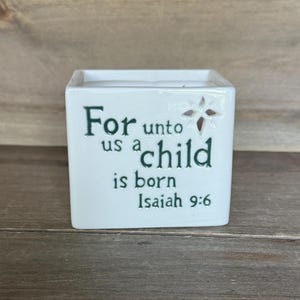 Puede incluir: Cubo de cerámica blanca con el texto "For unto us a child is born Isaiah 9:6" en verde oscuro. Un recorte en forma de estrella está en el lateral. El cubo está sobre una superficie de madera.