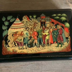 Puede incluir: Una caja de madera lacada en negro con una escena pintada de colores de un carruaje tirado por caballos, una tienda de campaña y personas con ropa tradicional rusa. La caja está decorada con ribetes dorados y tiene las palabras "Палех" y "Народные-сказки" escritas en la parte inferior.