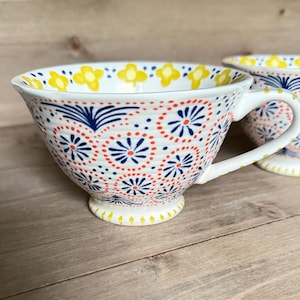 Op de afbeelding: Witte keramische theekop met een oor, versierd met een herhalend patroon van rode, blauwe en gele bloemmotieven. De theekop heeft een brede komvorm en staat op een kleine voet.