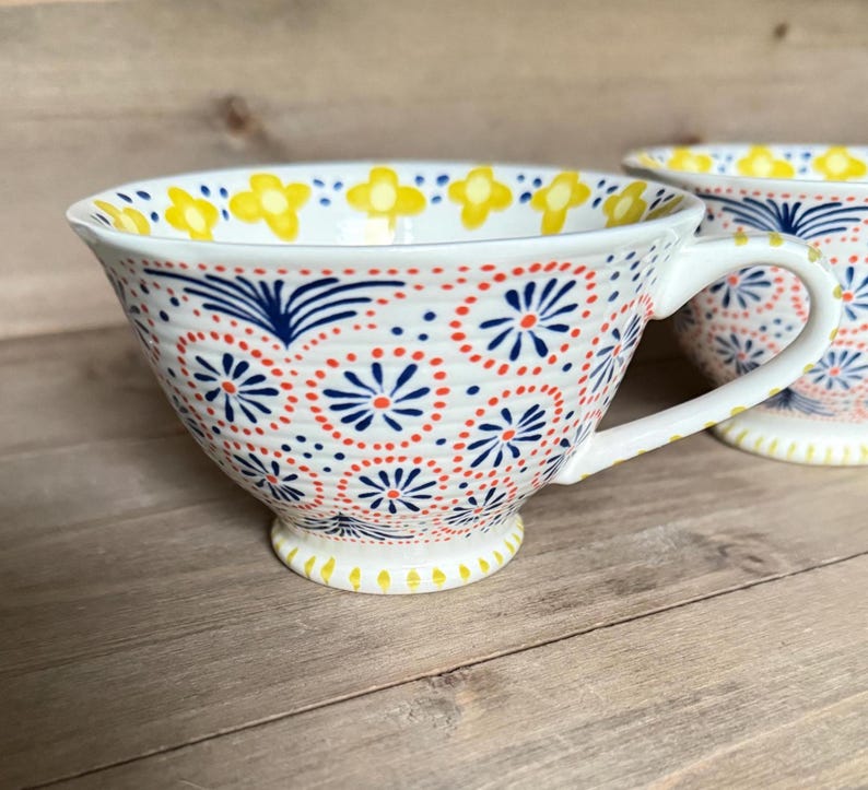 Op de afbeelding: Witte keramische theekop met een oor, versierd met een herhalend patroon van rode, blauwe en gele bloemmotieven. De theekop heeft een brede komvorm en staat op een kleine voet.