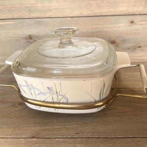 Pode incluir: Uma travessa quadrada vintage com tampa de vidro transparente e um design floral com íris roxas e verdes. A travessa está em um suporte de metal dourado com alças brancas. Uma peça clássica para assar e servir.