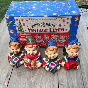 May include: Set of four vintage elf napkin rings. Each ring is hand-painted and features a different elf holding a different object. The elves are wearing red, green, and blue clothing. The box says "Christopher Radko Presents Shiny Brite Vintage Elves".