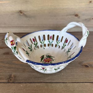 May include: A white ceramic basket with a blue rim and a braided handle. The basket features a pierced design and is decorated with hand-painted pink and red flowers and green leaves. The basket is approximately 25 cm in diameter.