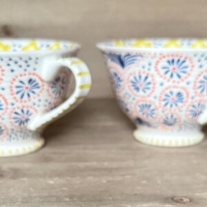 Op de afbeelding: Twee witte theekopjes met ingewikkelde bloempatronen in koraal, blauw en geel. De kopjes hebben een afgeronde vorm met een oor en staan op een kleine voet. Het ontwerp wordt op beide kopjes herhaald.