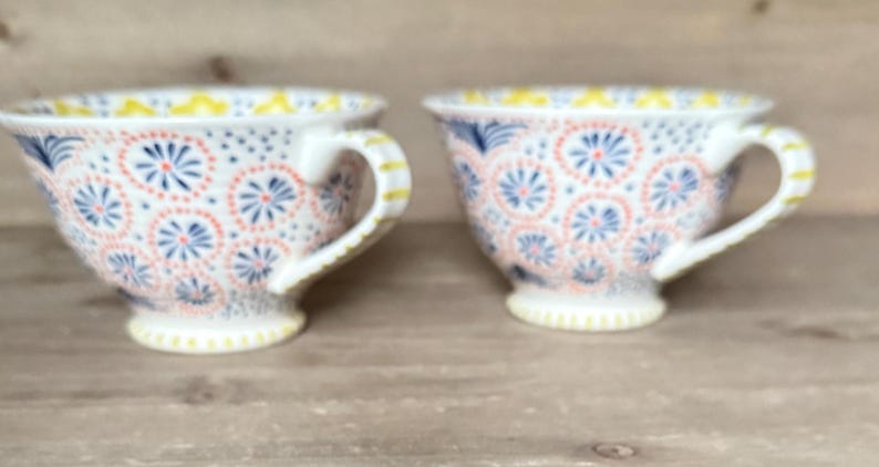 Op de afbeelding: Twee witte theekopjes met ingewikkelde bloempatronen in koraal, blauw en geel. De kopjes hebben een afgeronde vorm met een oor en staan op een kleine voet. Het ontwerp wordt op beide kopjes herhaald.