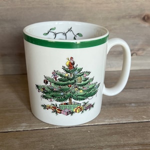 May include: White ceramic mug with a green rim and a Christmas tree design. The tree is decorated with ornaments, presents, and a Santa Claus figure at the top.