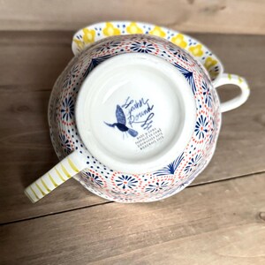Op de afbeelding: Een witte keramische theekop en schotelset met een kleurrijk bloemmotief. De kop heeft een handvat met gouden strepen en een blauw vogel logo aan de onderkant. Het ontwerp bevat rode, blauwe en gele accenten.