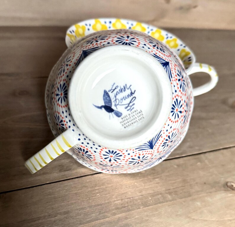 Op de afbeelding: Een witte keramische theekop en schotelset met een kleurrijk bloemmotief. De kop heeft een handvat met gouden strepen en een blauw vogel logo aan de onderkant. Het ontwerp bevat rode, blauwe en gele accenten.