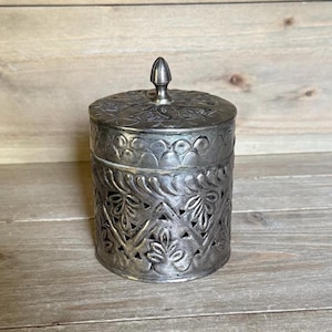 May include: A silver-colored, cylindrical container with a lid. The container features intricate floral and leaf cut-out designs. The lid is adorned with embossed patterns and a small, pointed finial. The container is likely made of metal.