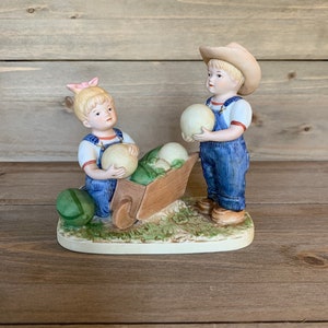 Può includere: Una statuetta in porcellana di due bambini, un ragazzo e una ragazza, vestiti con salopette, che tengono in mano grandi uova bianche. La ragazza sta spingendo un carretto pieno di altre uova. La statuetta è posta su una base verde erbosa.