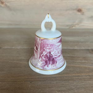 Pode incluir: Um sino de porcelana branca com uma borda dourada e uma cena de inverno rosa. O sino tem uma alça de laço no topo e uma cena de inverno rosa e branca com uma pessoa a cavalo.