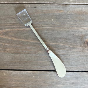 以下が含まれることがあります： 透明なプラスチックカバーが付いた銀色のバターナイフ。ナイフは丸みを帯びた刃と細長い柄をしています。透明なプラスチックカバーには円形のくぼみがあります。ナイフは木製の表面にあります。