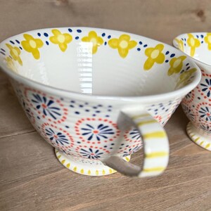 Op de afbeelding: Witte keramische theekop met een handvat, versierd met gele bloem- en rode en blauwe stippenpatronen. Het handvat heeft gele strepen. De theekop staat op een houten oppervlak.