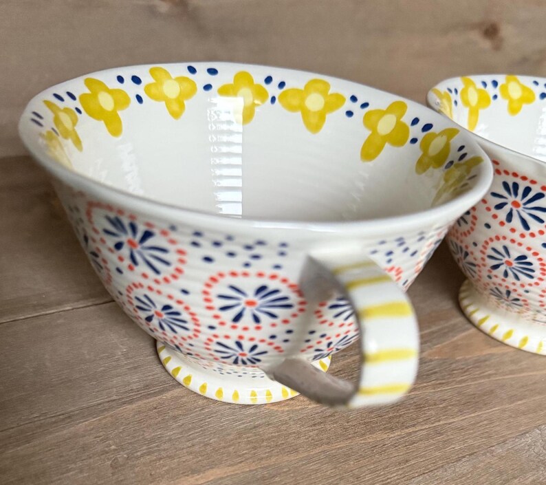 Op de afbeelding: Witte keramische theekop met een handvat, versierd met gele bloem- en rode en blauwe stippenpatronen. Het handvat heeft gele strepen. De theekop staat op een houten oppervlak.