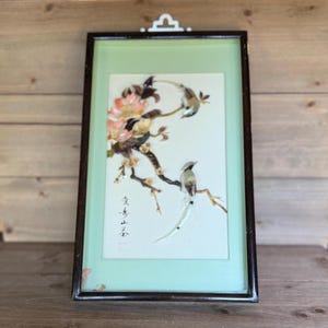 May include: Framed artwork featuring a detailed depiction of birds perched on flowering branches. The artwork has a light green background and is encased in a dark brown frame. The image includes Chinese characters.