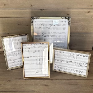 May include: Four picture frames with gold-colored metal frames. The frames hold sheet music. The frames are different sizes and shapes, including rectangular and square. The frames are arranged on a wooden surface.