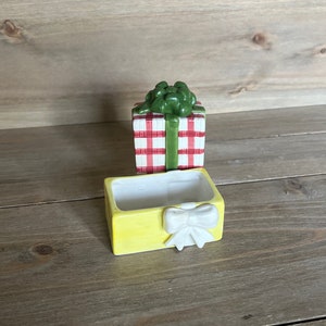 May include: A ceramic gift box with a red and white checkered pattern and a green bow. The box is open and the inside is yellow with a white bow.