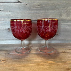 Puede incluir: Dos copas de vino rojo rubí con bordes dorados. Las copas tienen un tallo y una base transparentes. El cuenco de cada copa está grabado con un diseño de vid. Las copas se exhiben sobre una superficie de madera.