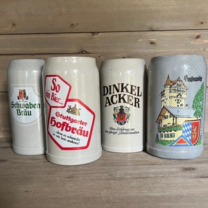 May include: Four ceramic beer steins with various logos and text. The steins are off-white and gray, with designs including the words "Schwaben Bräu", "Stuttgarter Hofbräu", and "Dinkel Acker". The steins are displayed on a wooden surface.