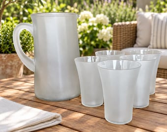 May include: A set of frosted glass drinkware consisting of one large pitcher with a curved handle and four matching tumblers. The glassware features a smooth, semi-opaque white finish and a simple, cylindrical design for serving beverages.